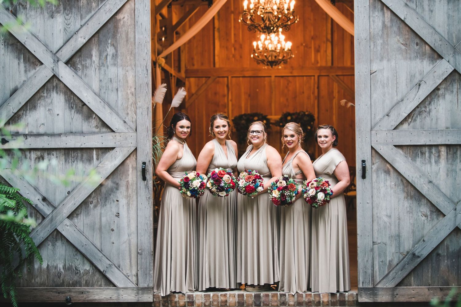 Wedding party poses in matching champagne dresses in front of a rustic barn door with chandeliers.
