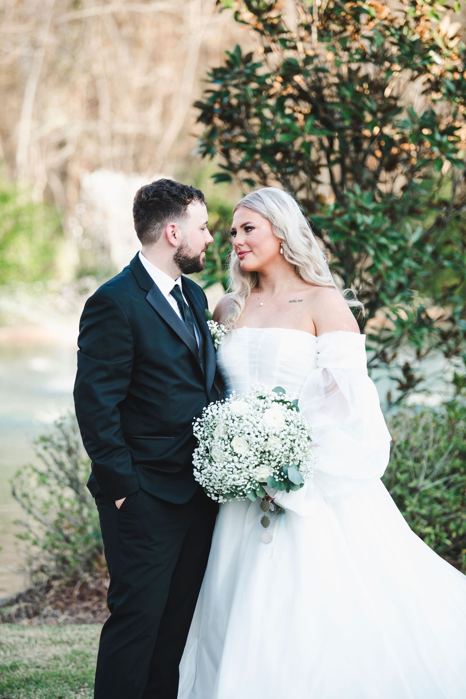 Elegant wedding couple poses with white bouquet against garden backdrop.