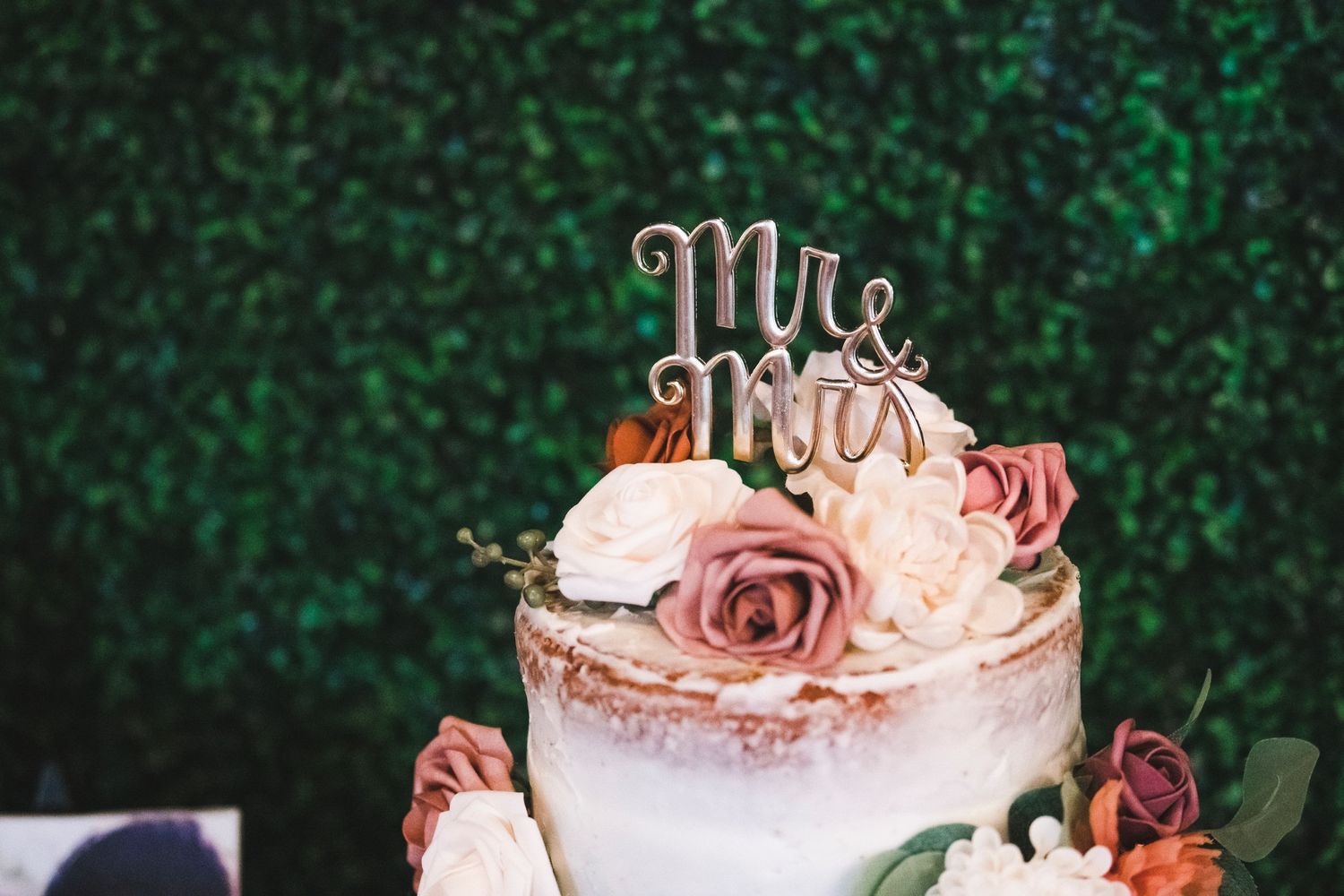 Elegant wedding cake decorated with pink roses and a 'Mr & Mrs' topper against a green foliage background.