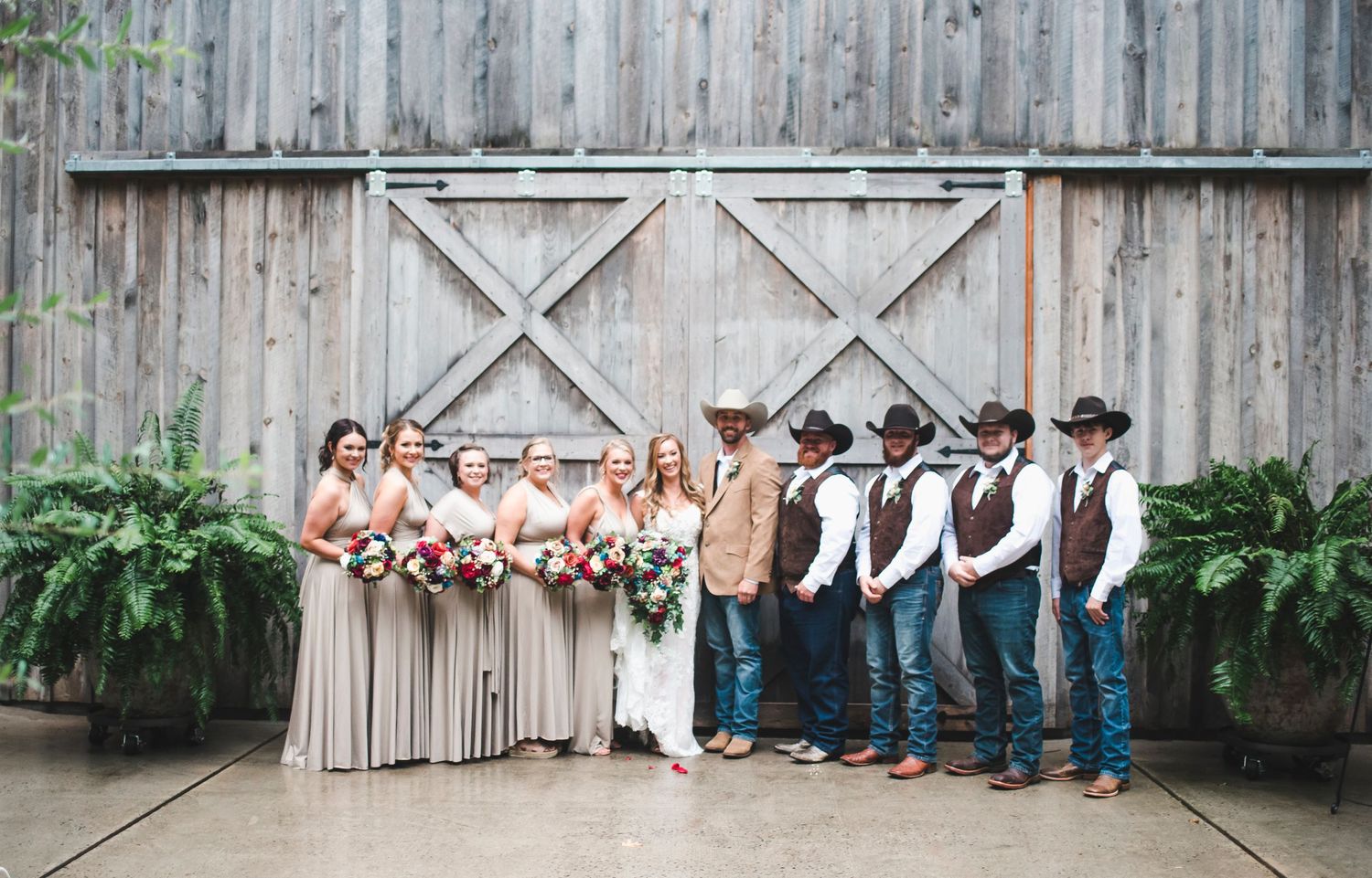 Wedding party in cowboy-western attire poses against weathered barn wall outdoors.