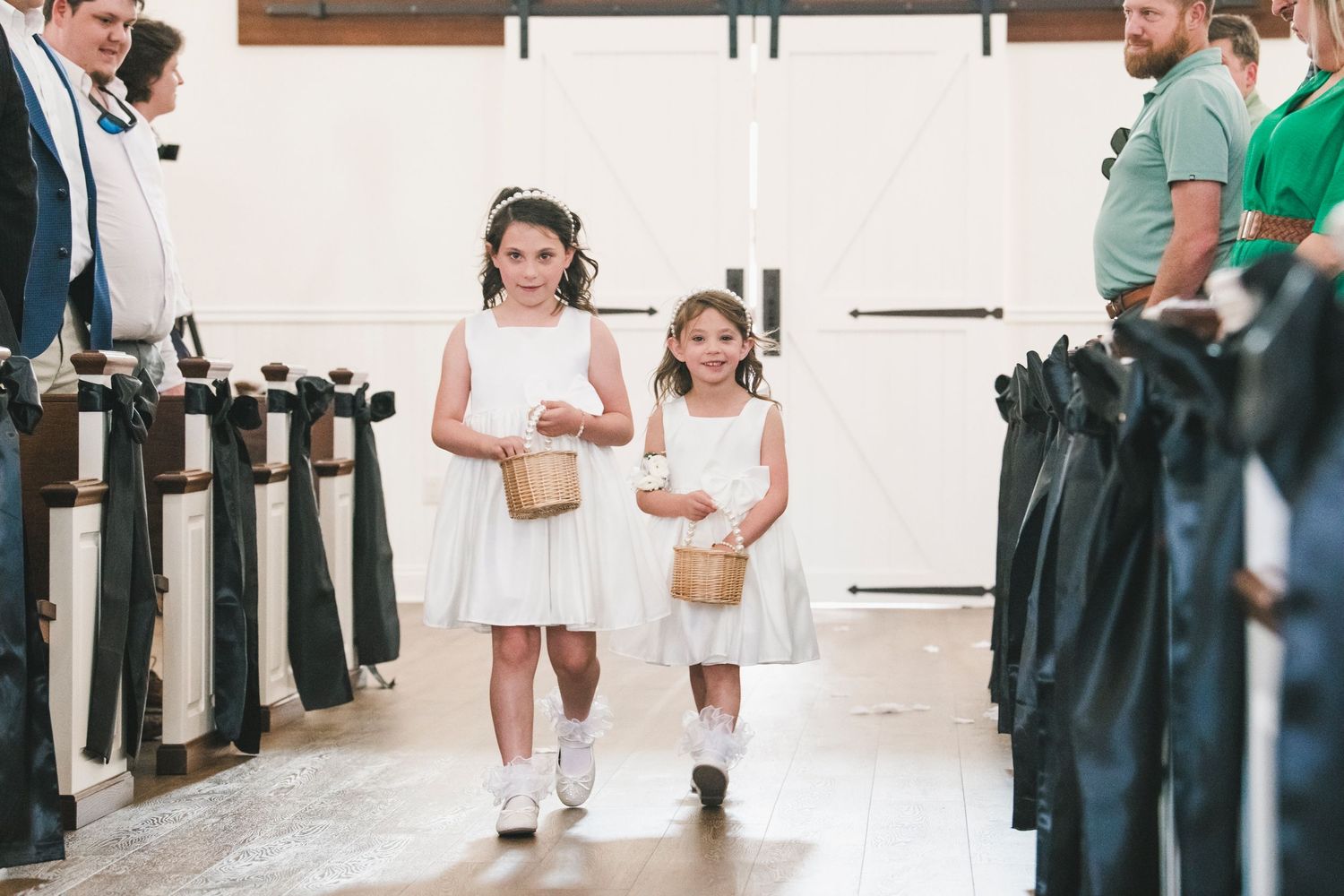 Two flower girls in white dresses walking down the aisle during a wedding ceremony.