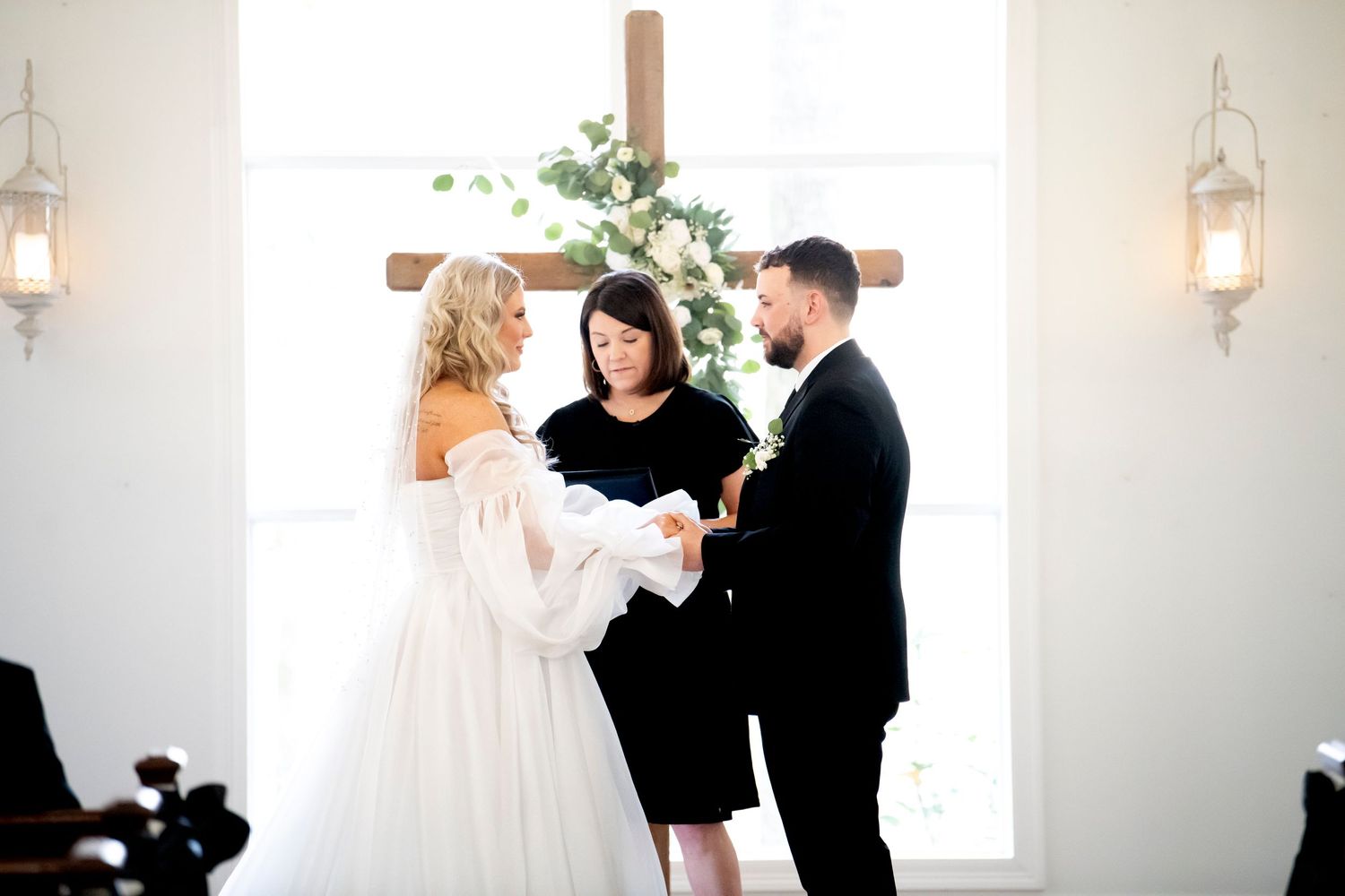 A wedding ceremony in progress inside a white chapel with natural light streaming through windows.