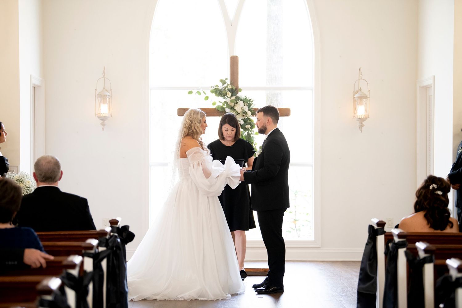 An intimate wedding ceremony in a bright white chapel with wooden pews and wall sconces.