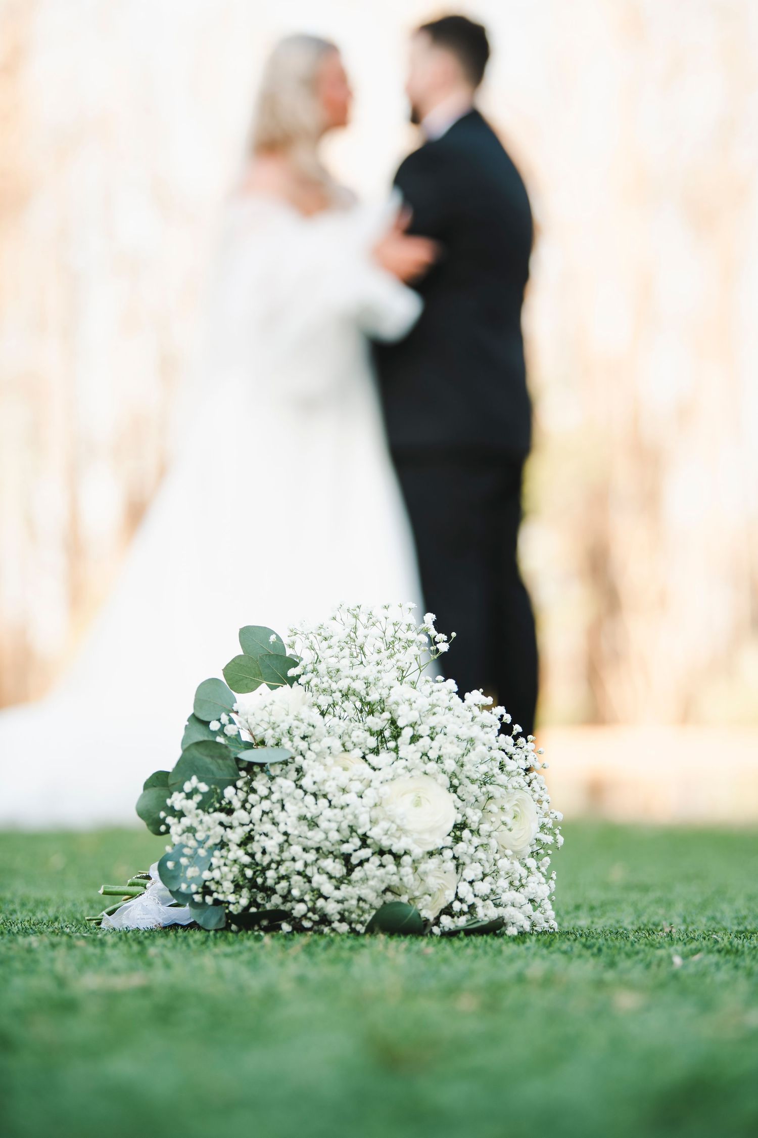 Delicate white and baby's breath bouquet on grass with blurred couple in background.