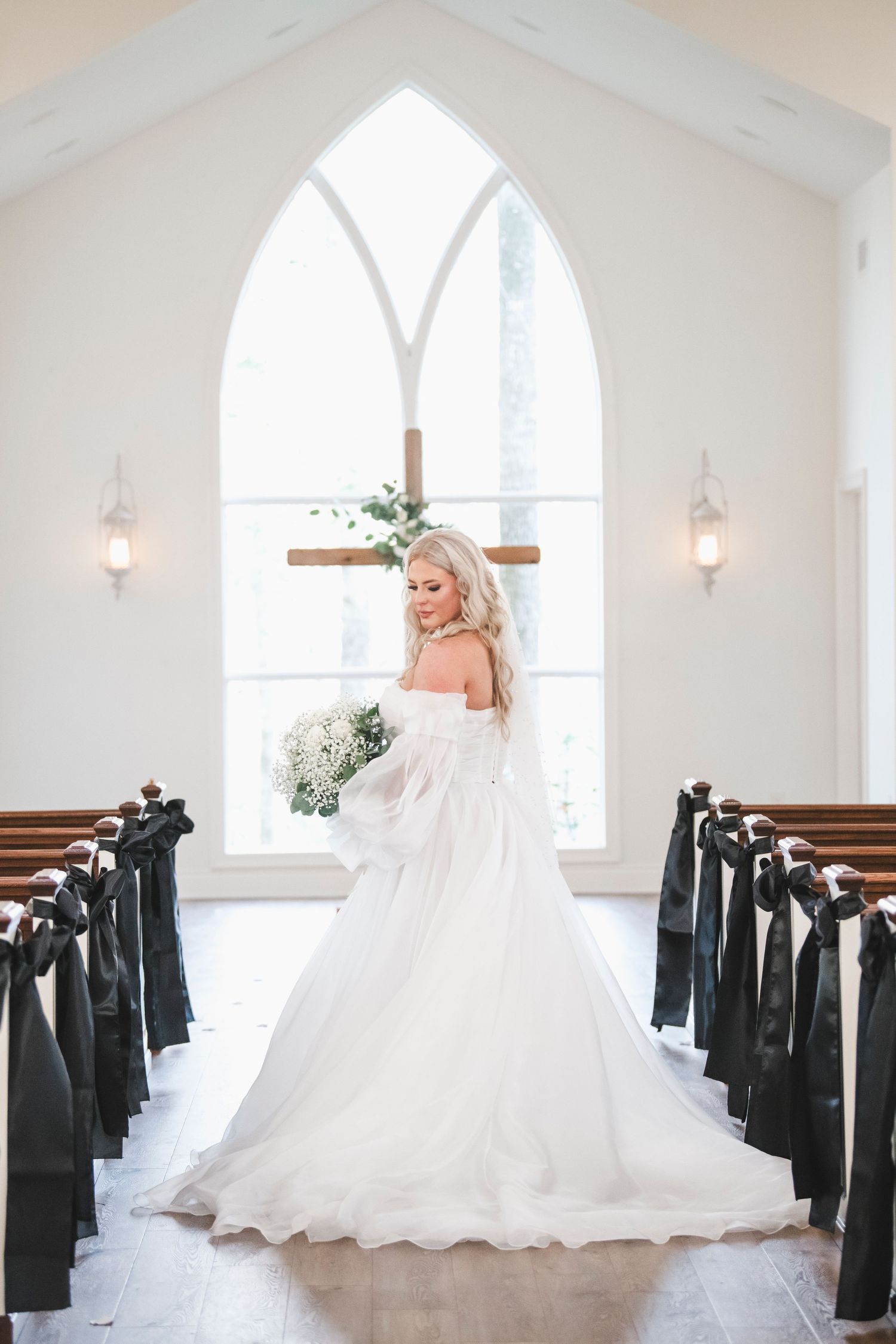 Bride in white ballgown poses in church aisle with Gothic arched window and pews decorated with black bows.