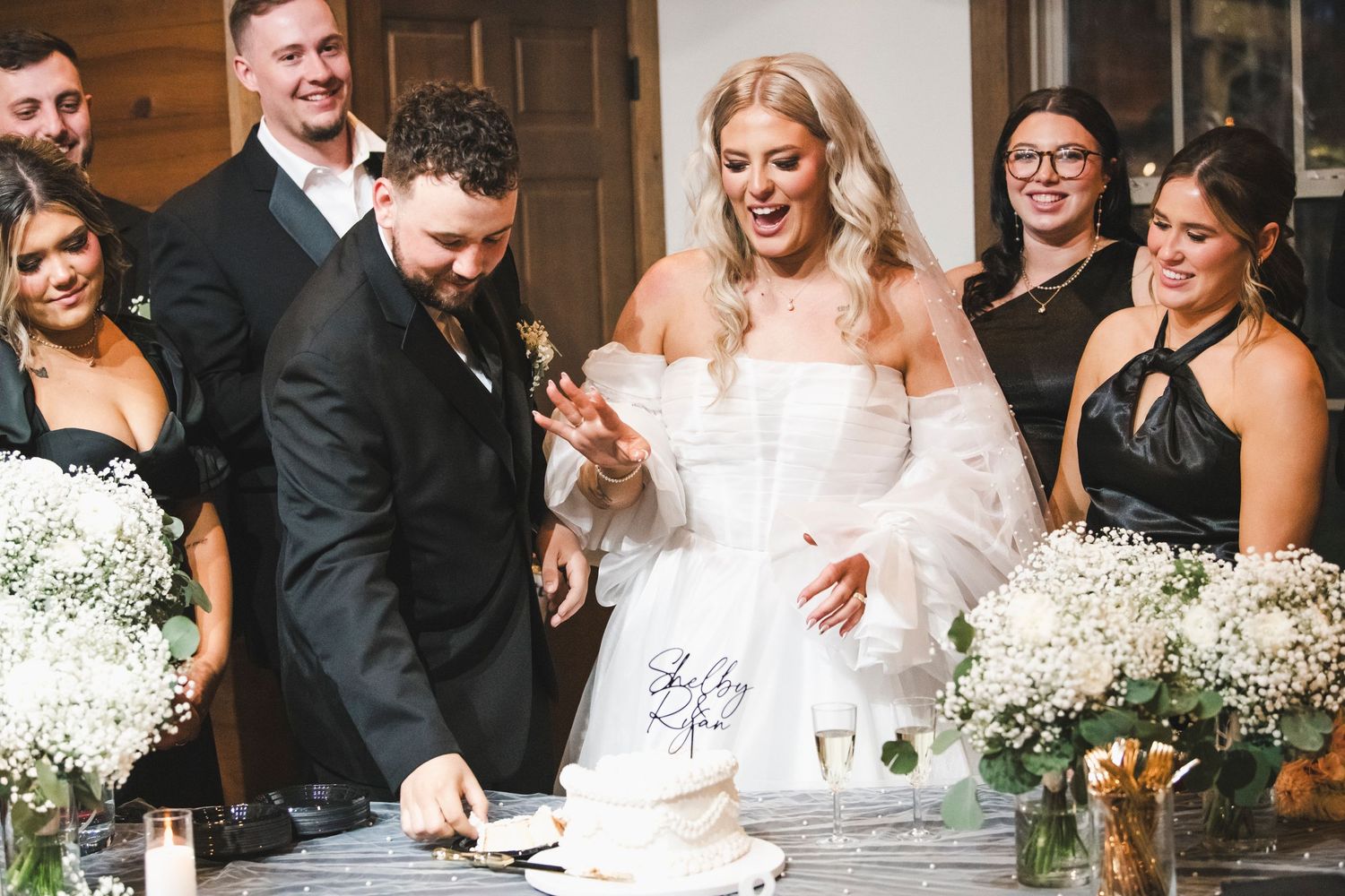 Wedding party gathers around a cake table as bride in white dress celebrates a special moment.