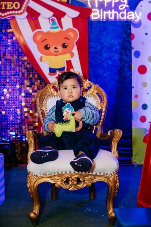 Small child sitting on ornate gold throne chair against carnival-themed backdrop.