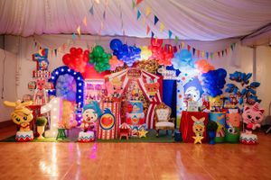 Colorful circus-themed birthday party decoration setup with balloons, banners and carnival props against a draped white backdrop.