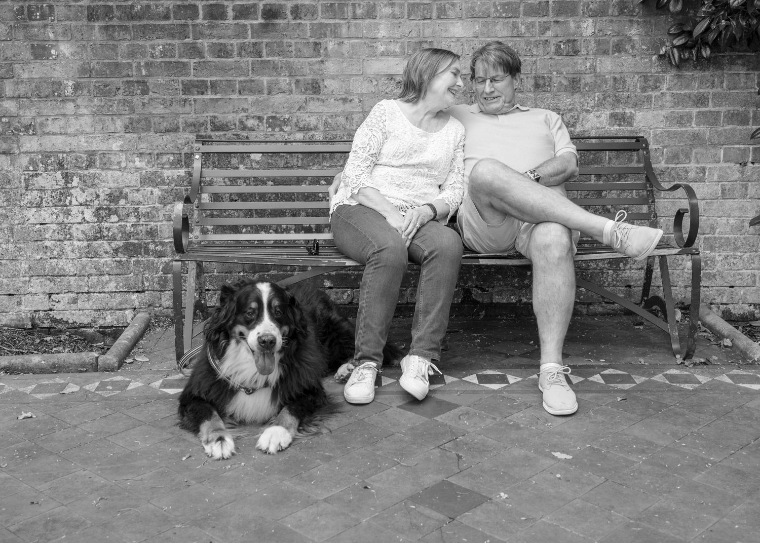A couple relaxes on a wooden bench beside a brick wall with their Bernese Mountain Dog lying nearby.