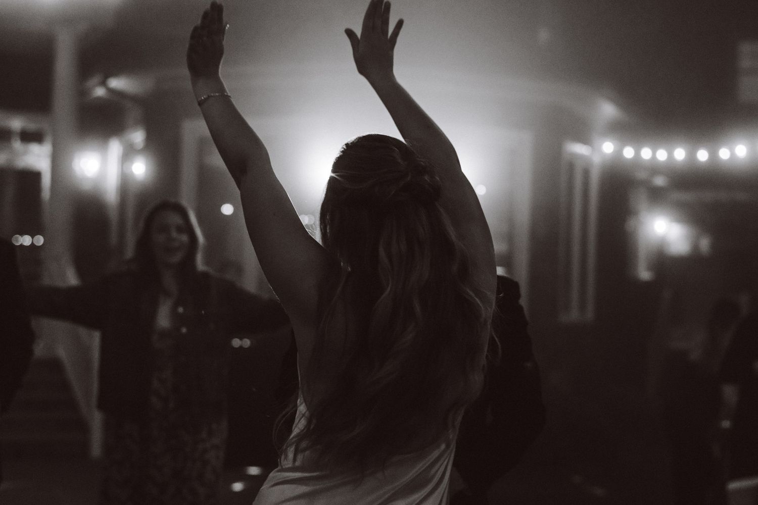 Black and white photo of bride dancing with arms raised at festive reception with string lights.