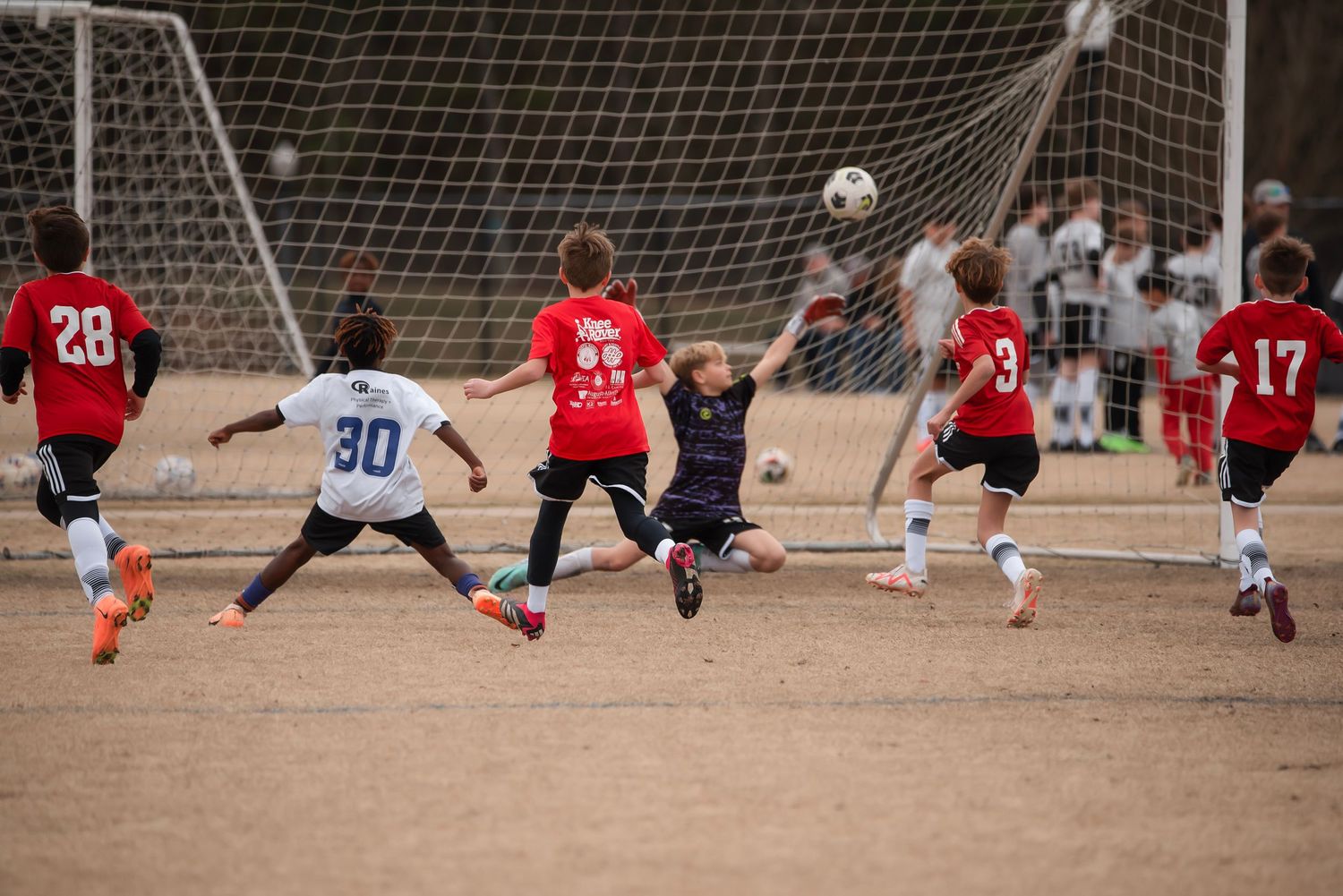 Dramatic soccer goal attempt with diving players and goalie at net.