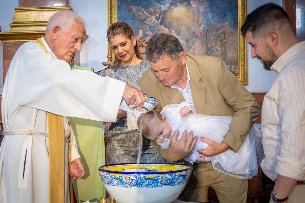 Priest performing Catholic baptism ceremony, pouring holy water over infant held by godfather over ornate baptismal font.
