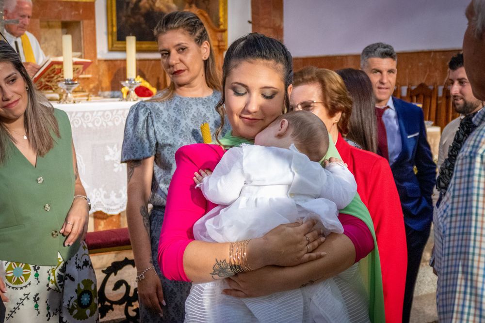Young woman in pink dress tenderly holds sleeping baby in white christening gown inside a church.