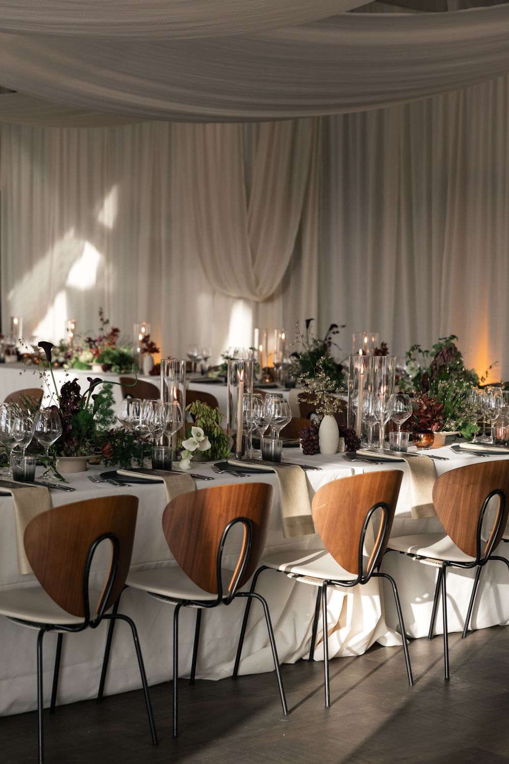 Modern wedding table setting with wooden chairs and floral centerpieces under white draped ceiling.