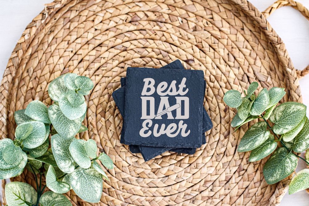 Navy blue coaster with 'Best Dad Ever' text displayed on woven straw placemat with decorative green leaves.