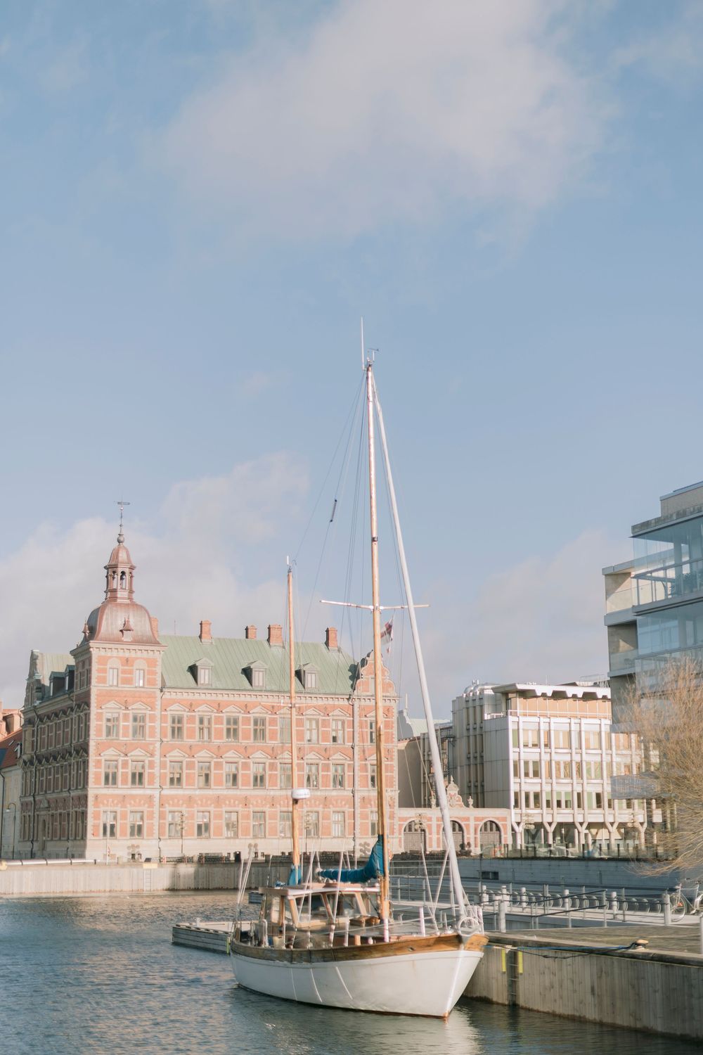 Sailboat docked in urban harbor with historic red brick building and blue sky.
