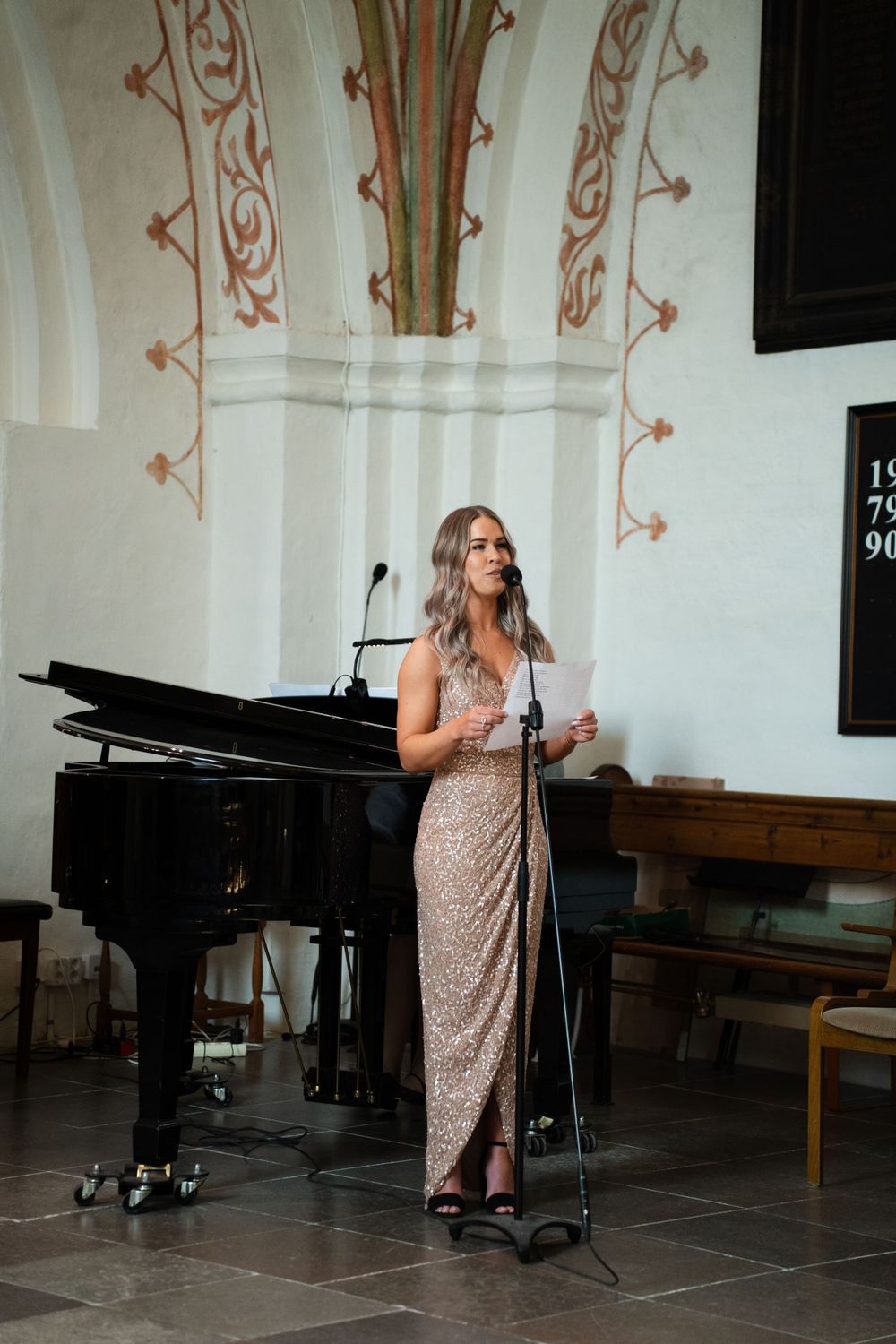 Performer in sequined gown stands next to grand piano in church with decorative walls.