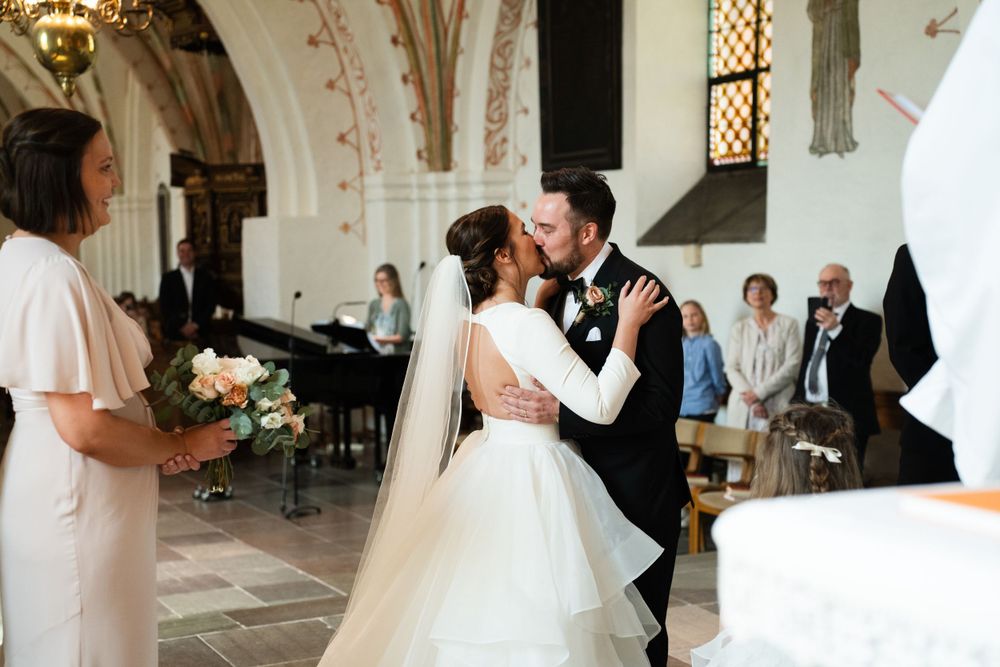 A couple shares their first kiss during a wedding ceremony in an ornate church with vaulted ceilings and stained glass.
