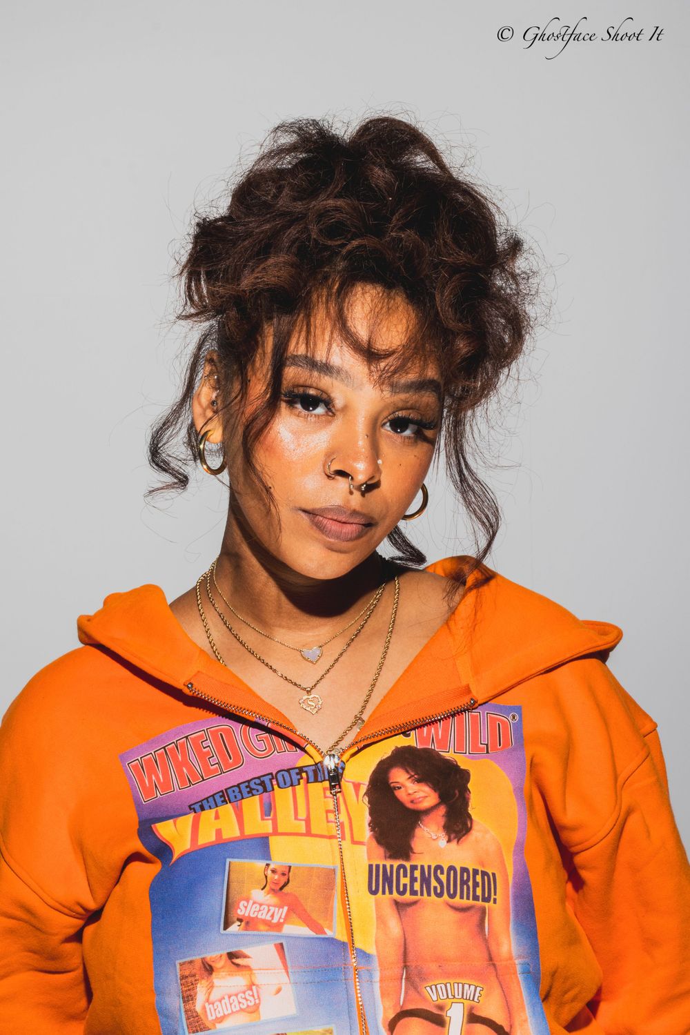 Portrait of person in orange hoodie with curly updo hairstyle against gray background.