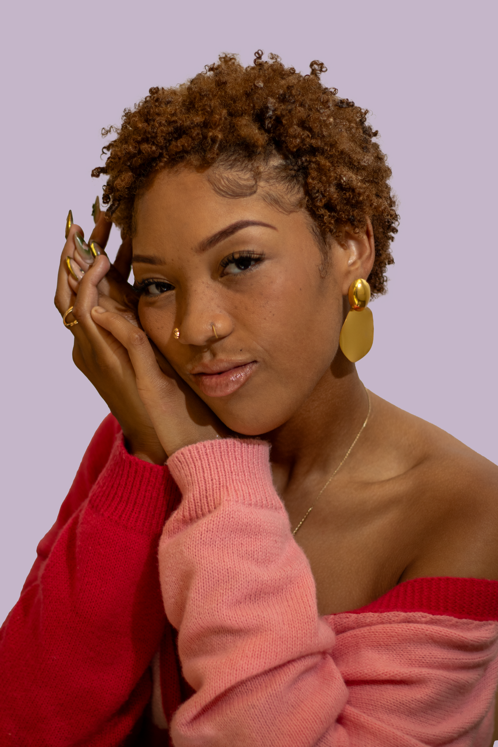 Casual portrait featuring natural curly hair, gold hoop earrings, and pink off-shoulder sweater against purple background.