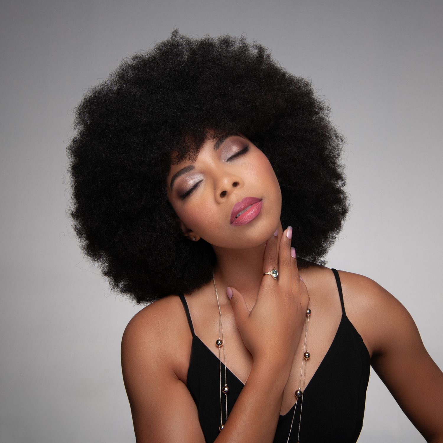 Portrait against gray backdrop featuring natural afro hairstyle, black top, and closed eyes in glamorous pose.