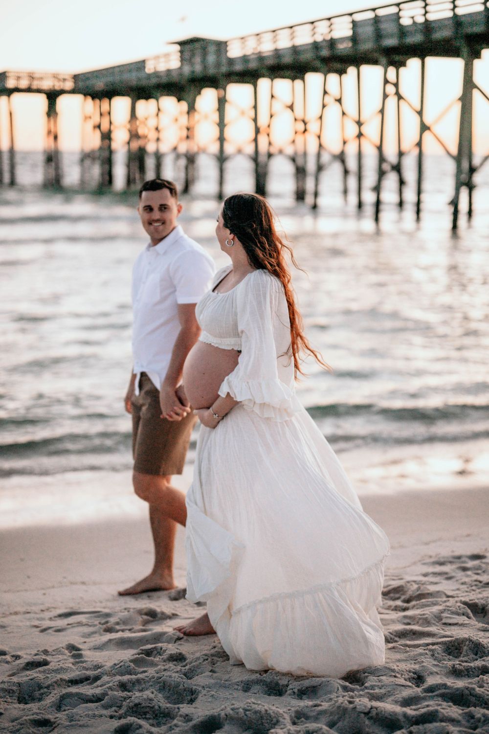 Couple walking along beach at sunset near a wooden pier.
