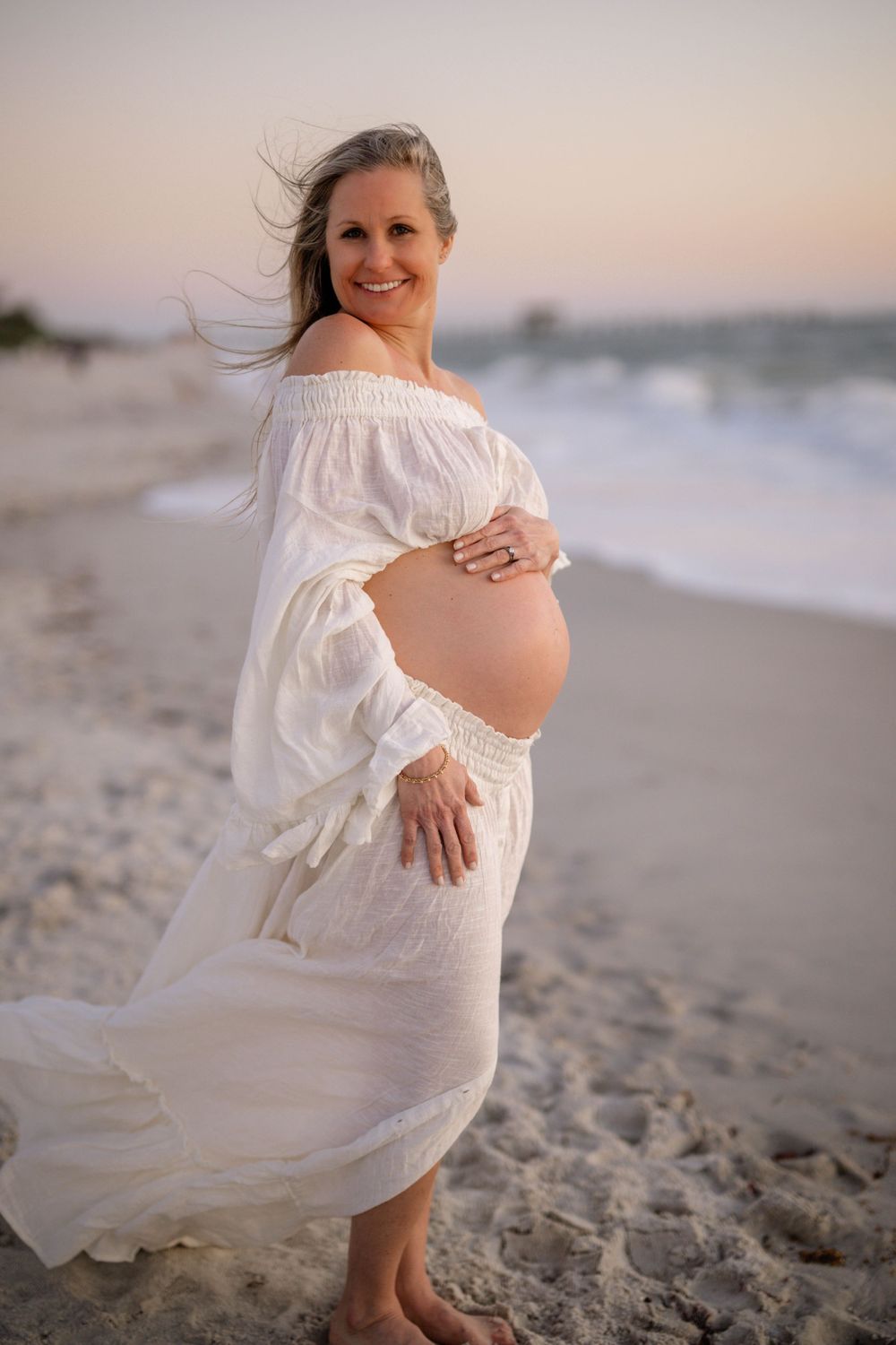 Expectant mother in flowing white dress poses on beach at sunset.