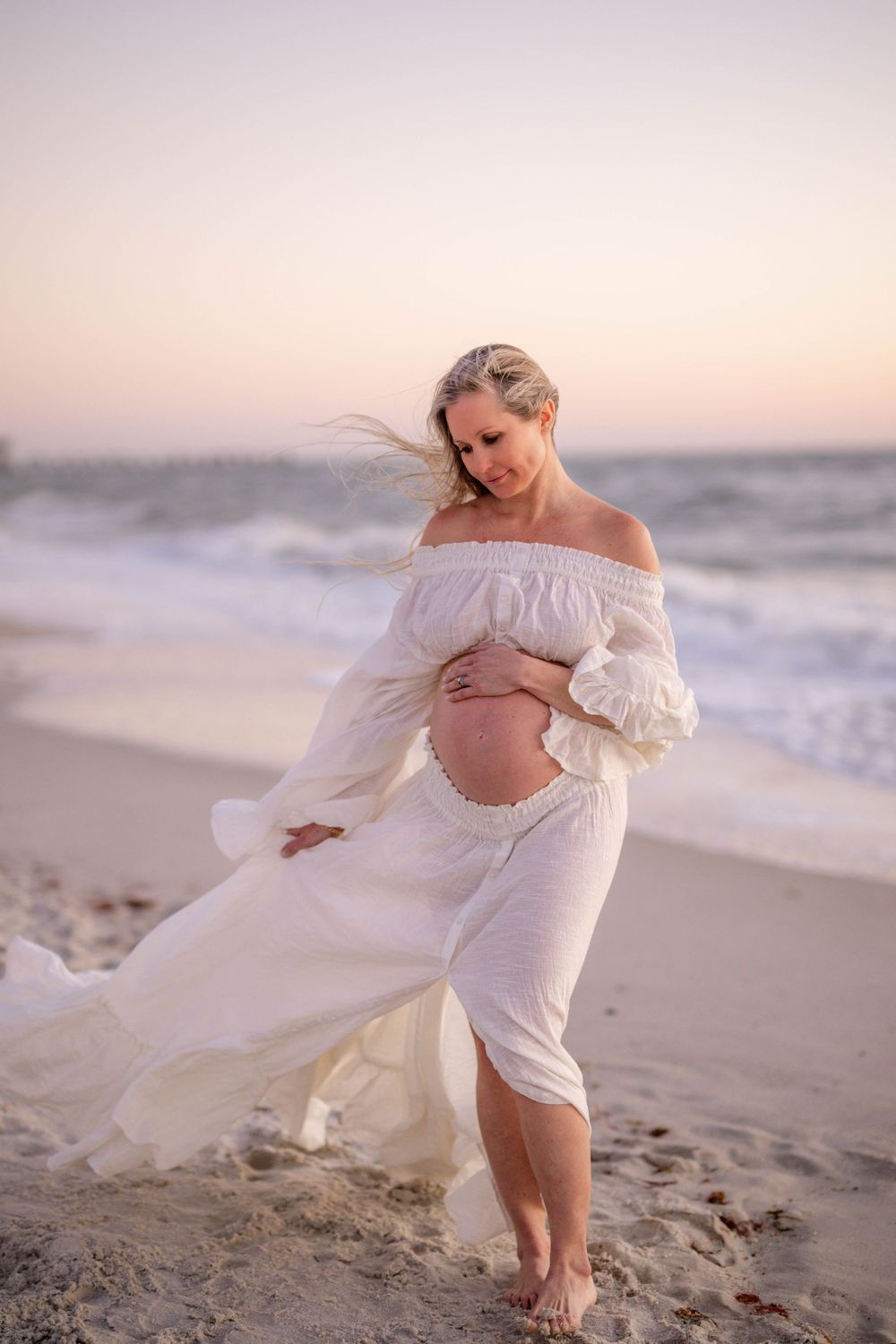Wind-swept maternity photo on beach with flowing white dress at dusk.