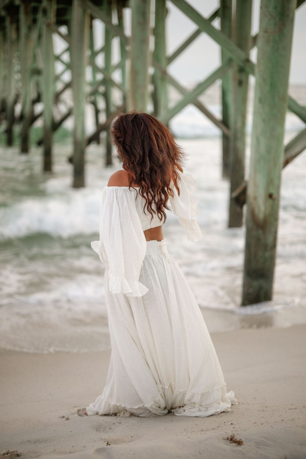A billowing white dress catches the breeze beneath a beach pier during a seaside photoshoot.