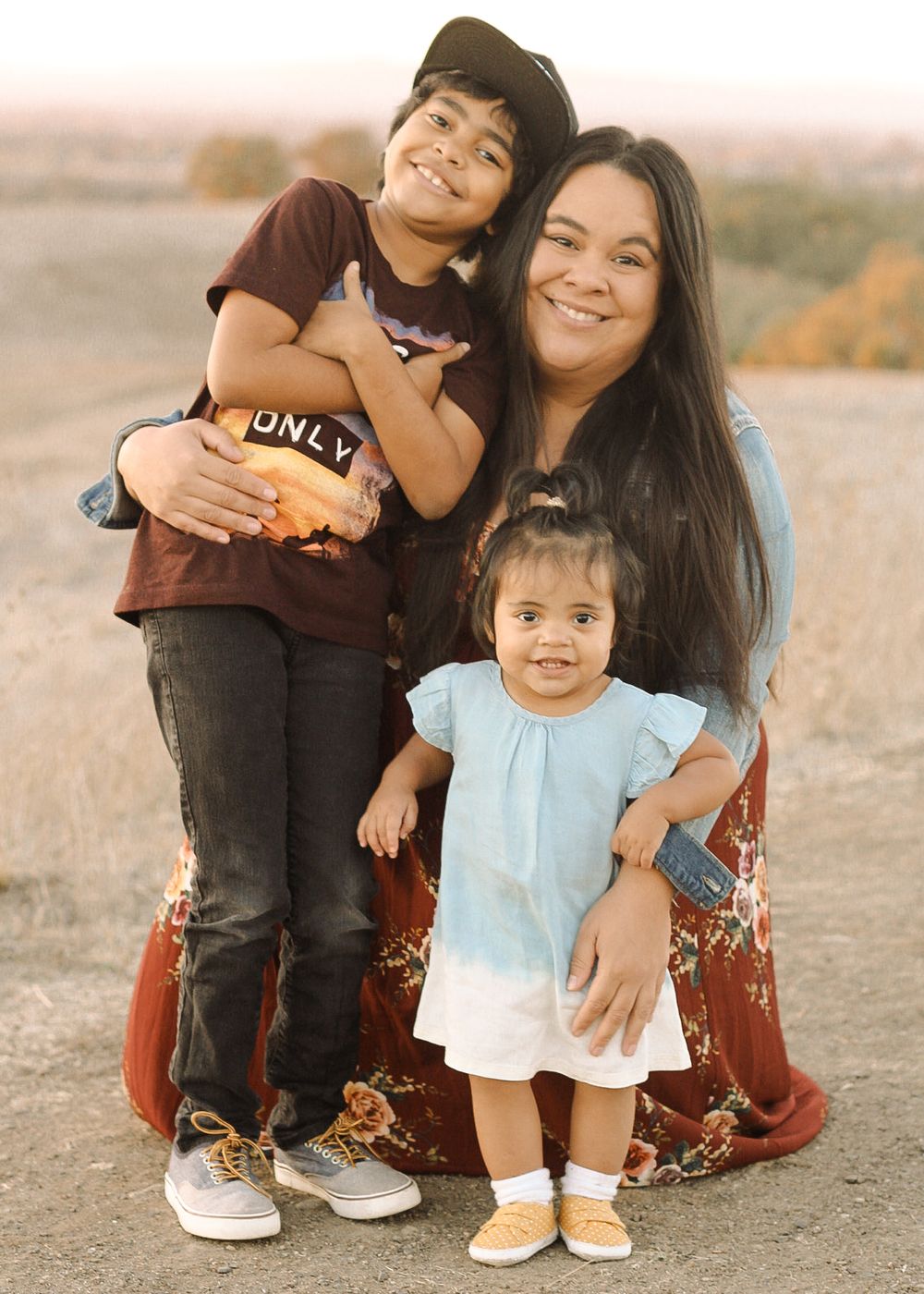 Mother loosely embracing son and daughter, all smiling and joyful, grassy hills in background for family session.
