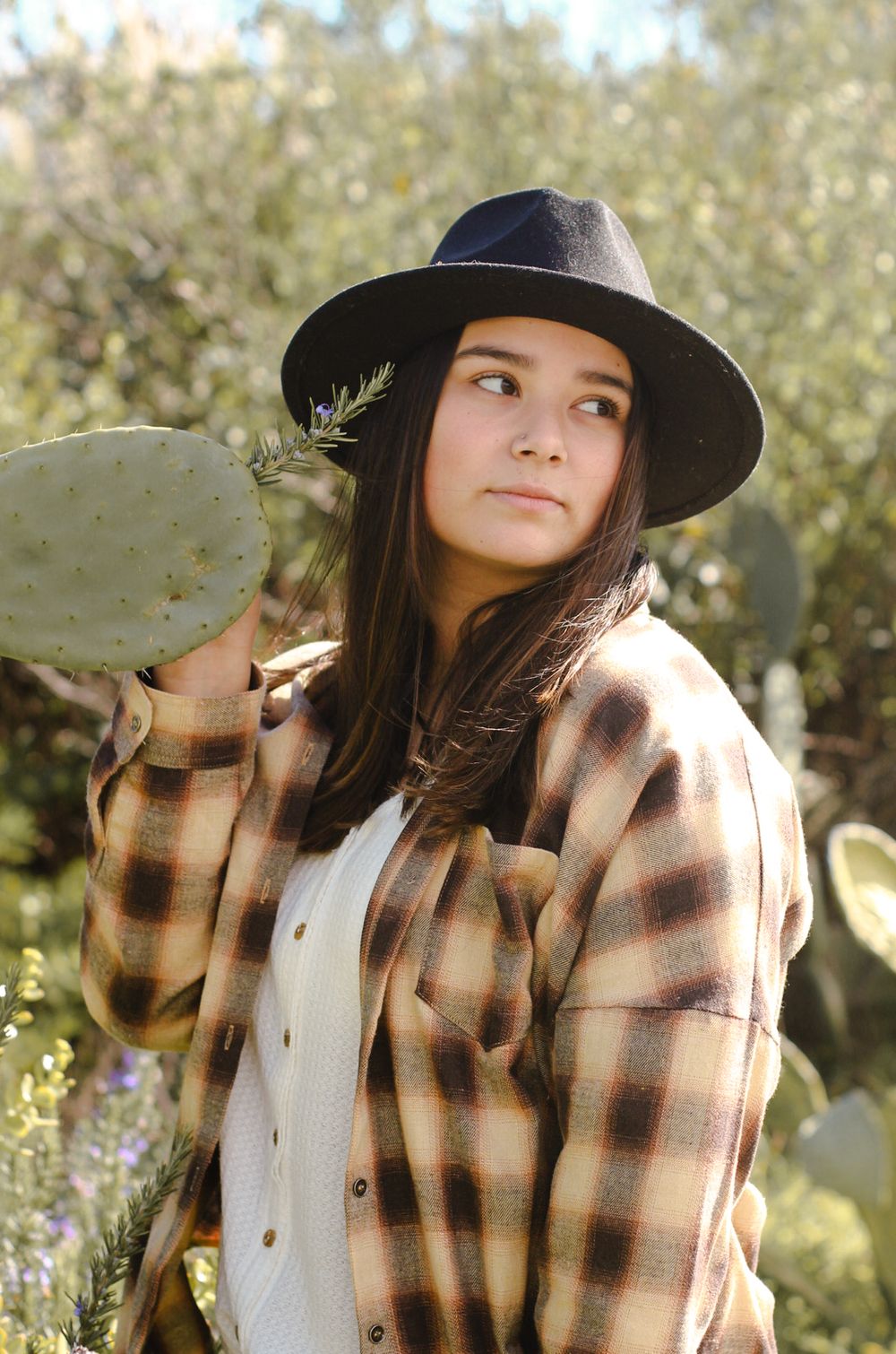 Woman in black hat and yellow and black checkered flannel posed looking away in front of plants including a cactus.