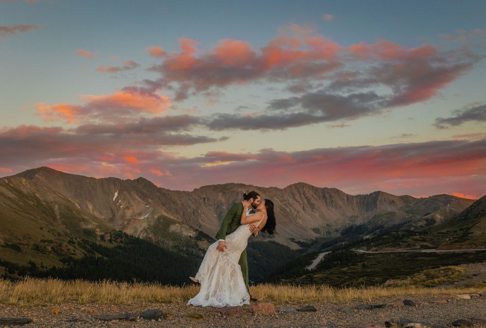 A romantic wedding portrait against a dramatic mountain sunset with pink and orange clouds.
