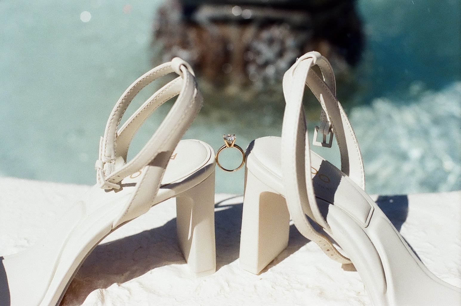 White bridal heels with a gold engagement ring placed between them on a sunny white surface.