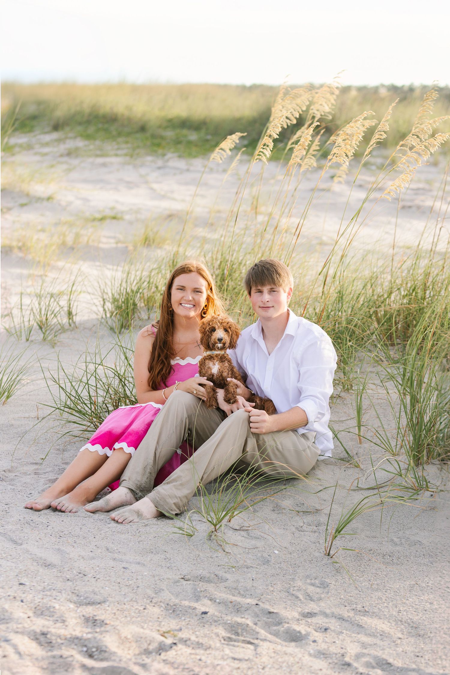 Couple relaxes on sandy beach with their goldendoodle puppy among sea oats at sunset.