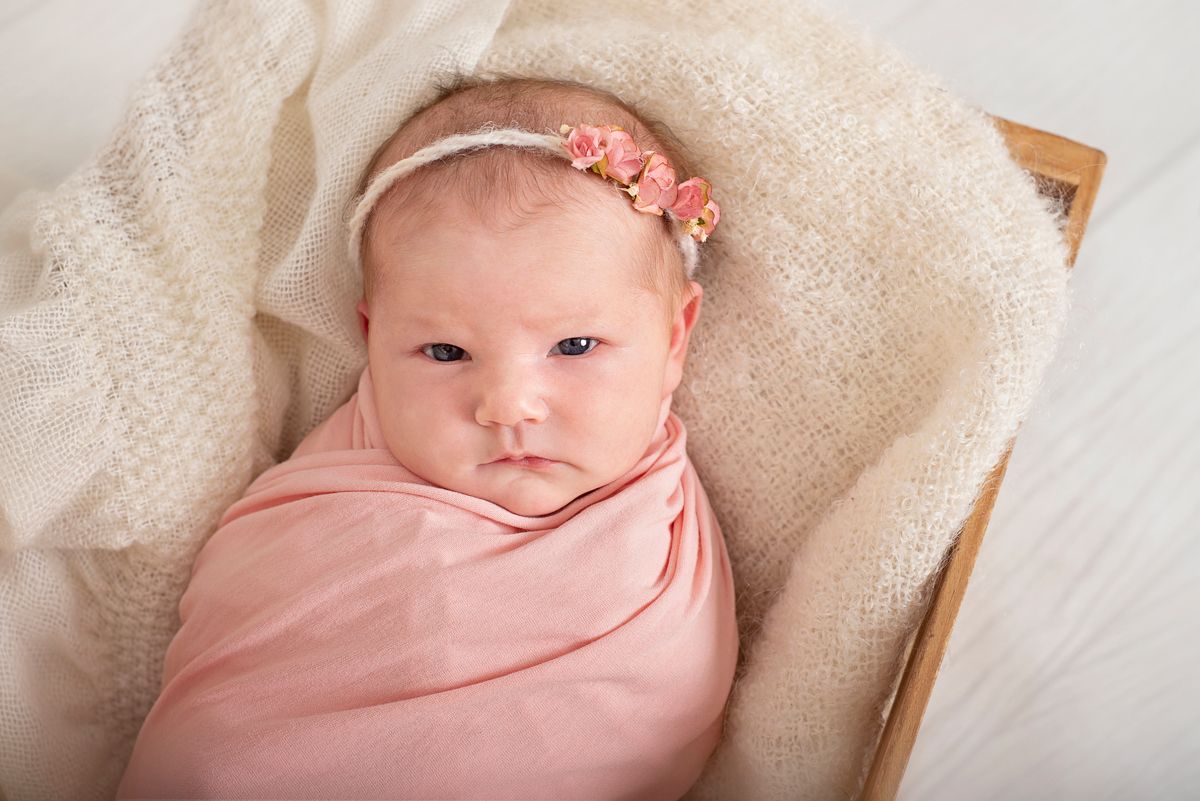 Awake newborn baby girl in wooden prop with white and pink blankets and delicate headband