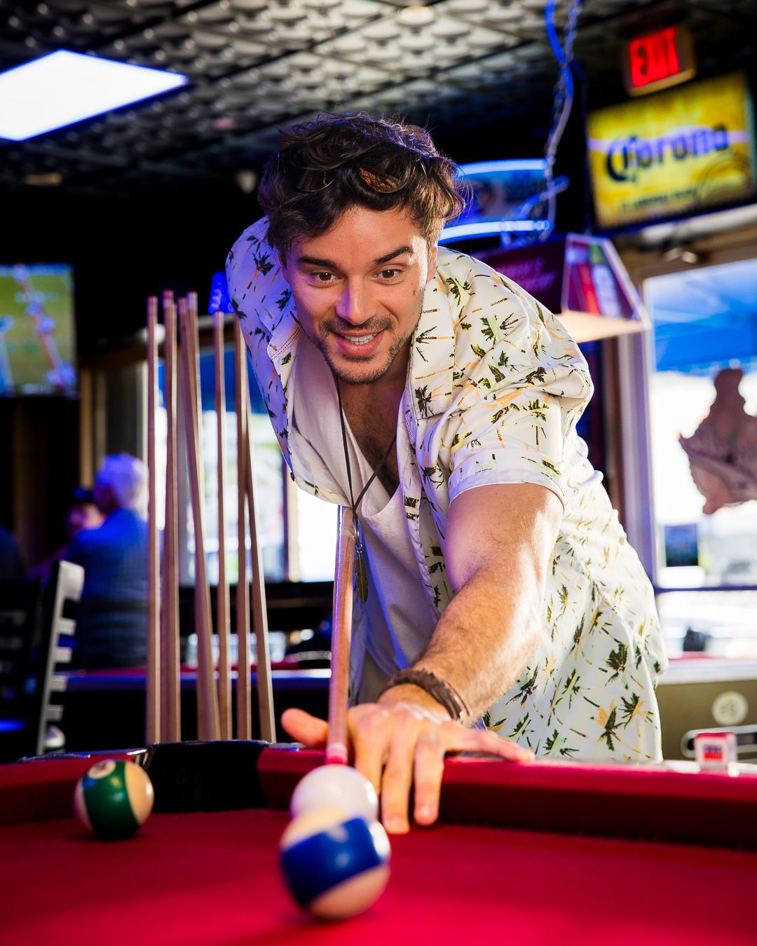 Someone in a floral shirt leans over a pool table about to take a shot in a bar with blue lighting and Corona signs.