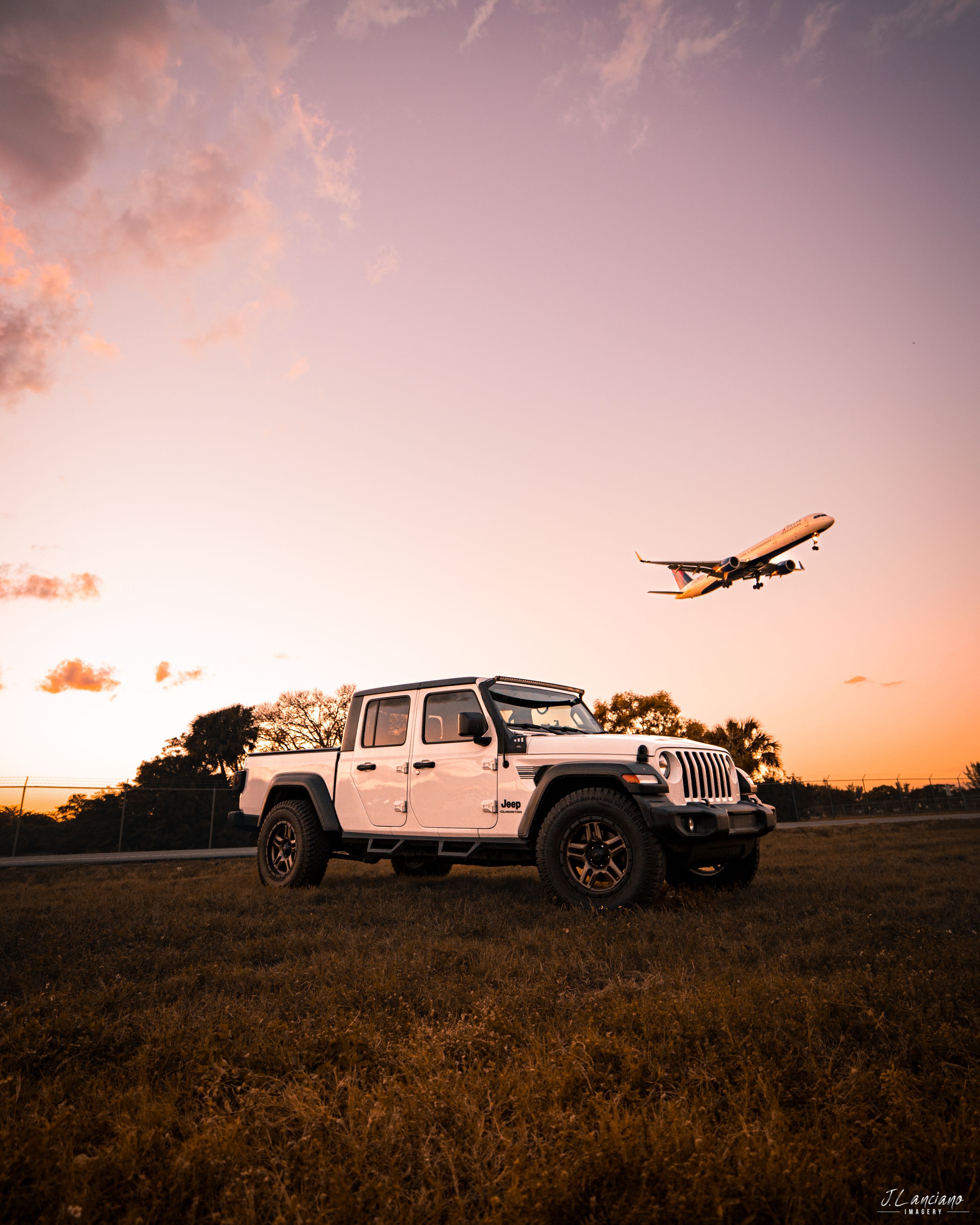 White Jeep Gladiator parked in field with passenger plane silhouetted against sunset sky.