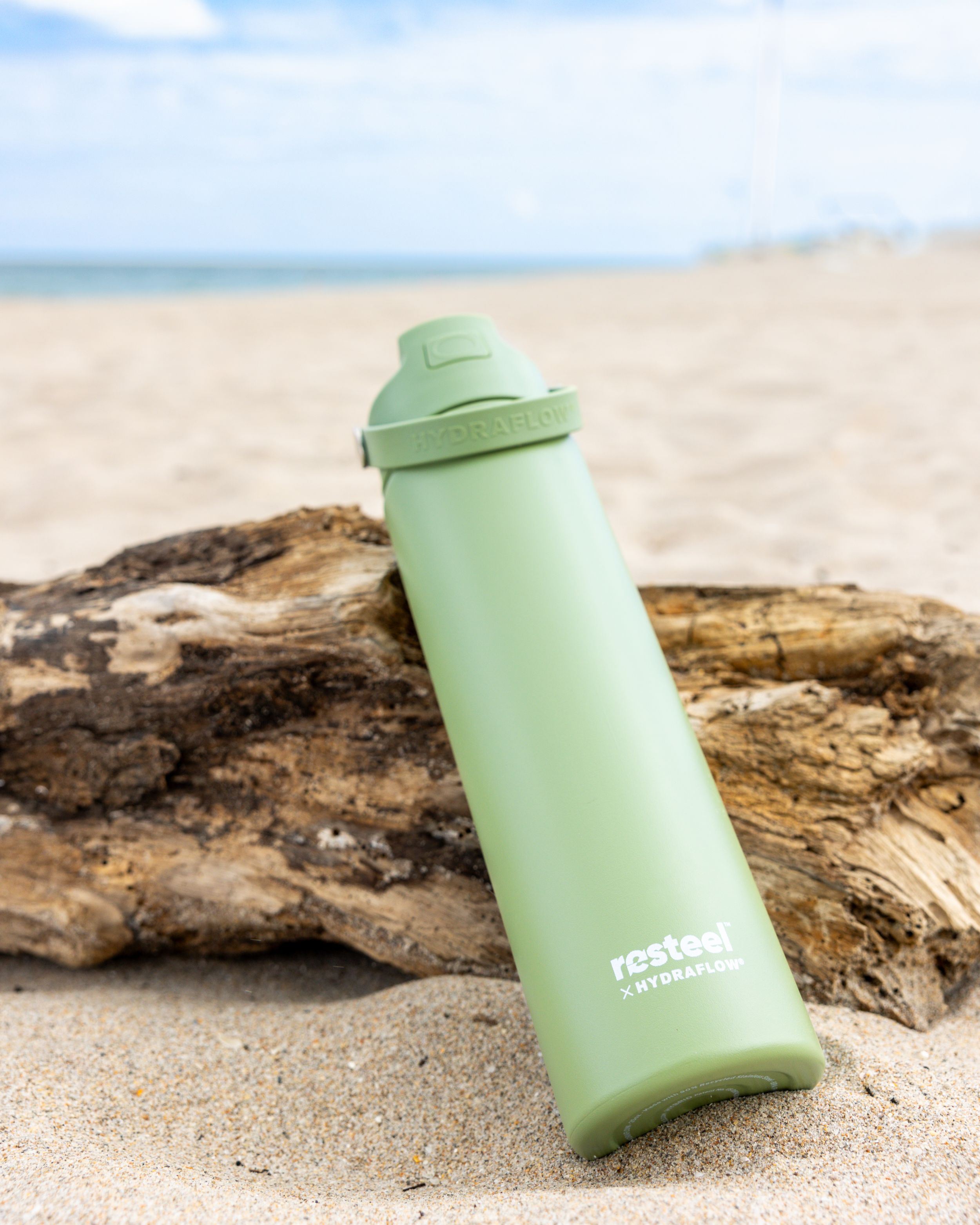 Mint green reusable water bottle sitting on driftwood at the beach with ocean in background.