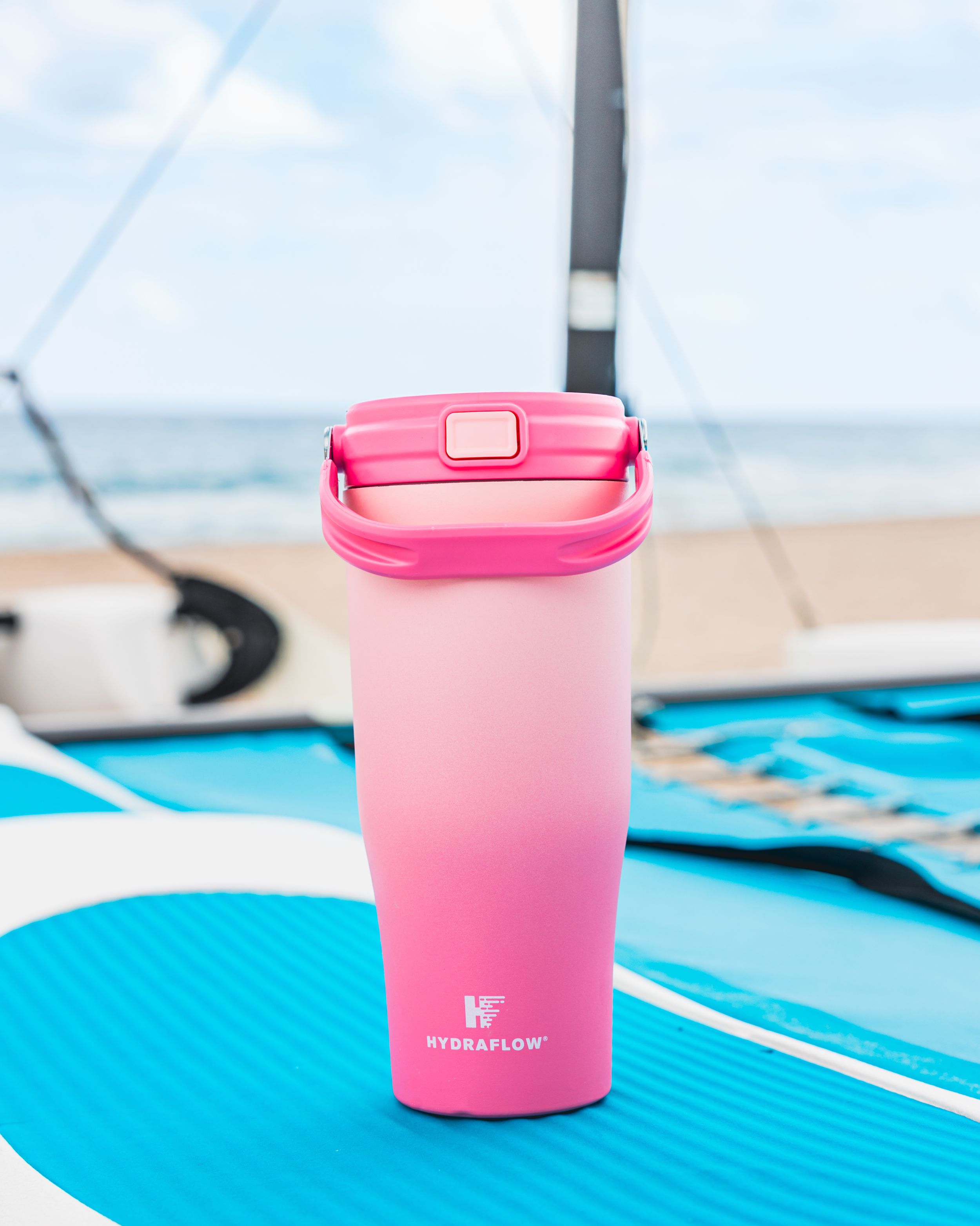Pink ombre travel tumbler on blue surfboard at beach with ocean in background.