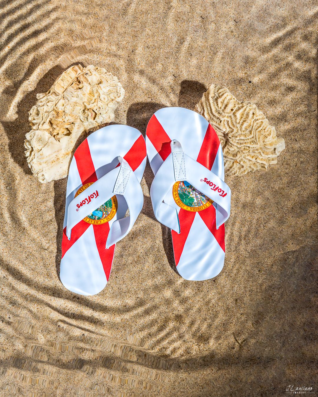 Red and white patterned flip flops on rippled beach sand with watery reflections.