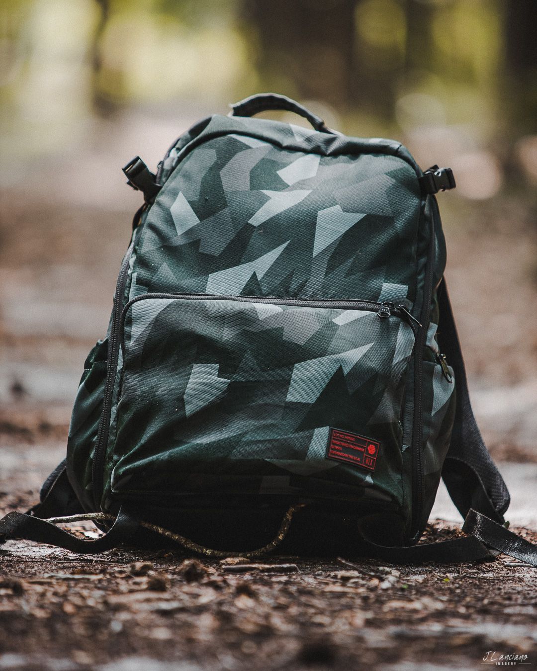 Camouflage patterned backpack with red label sitting on forest ground covered in leaves.