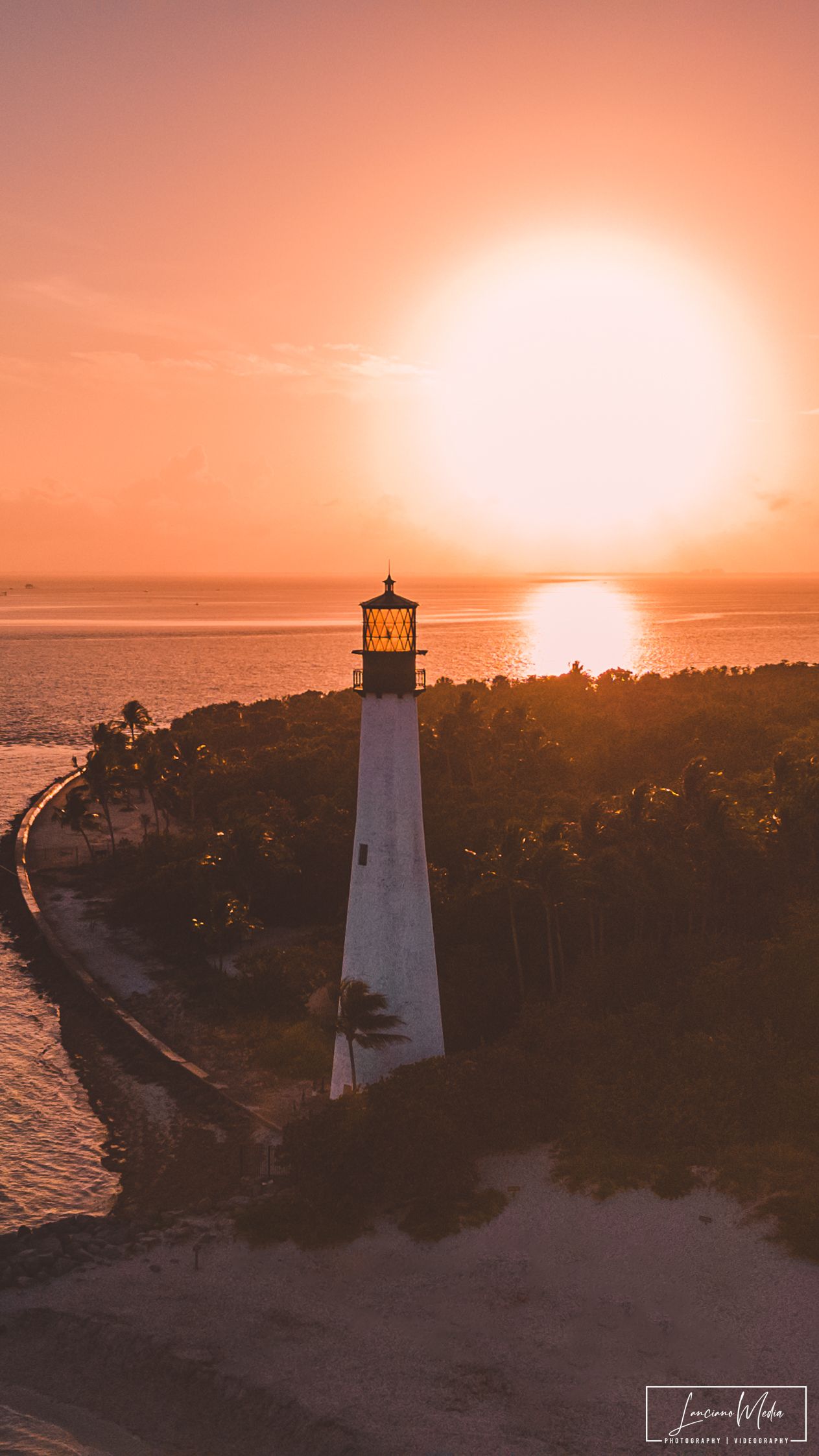 White lighthouse silhouetted against dramatic orange sunset over coastal waters.