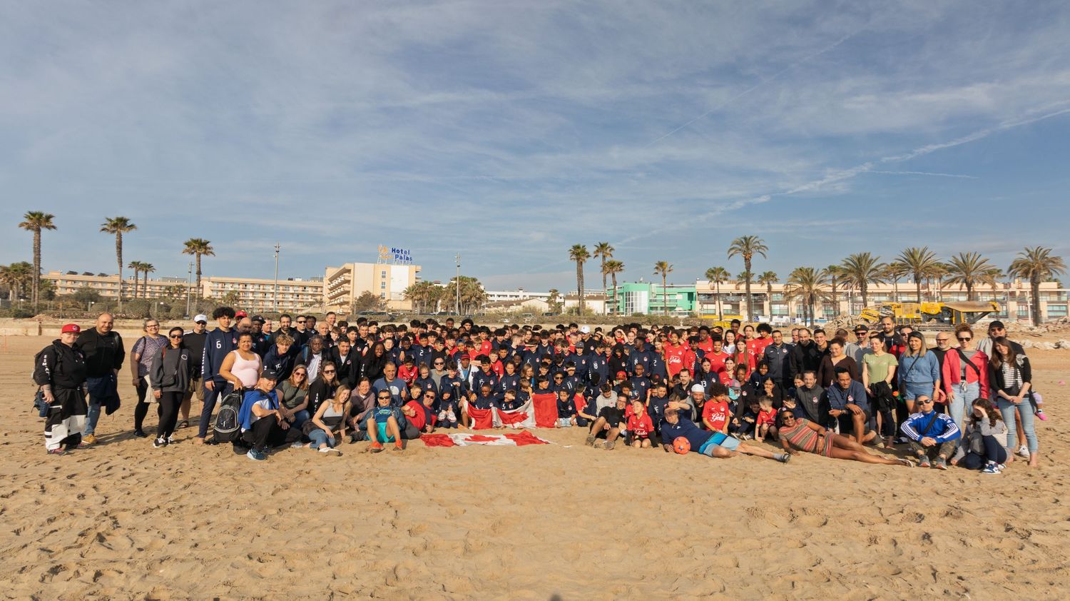 Large group photo of soccer players and fans gathered on a sunny beach with palm trees.