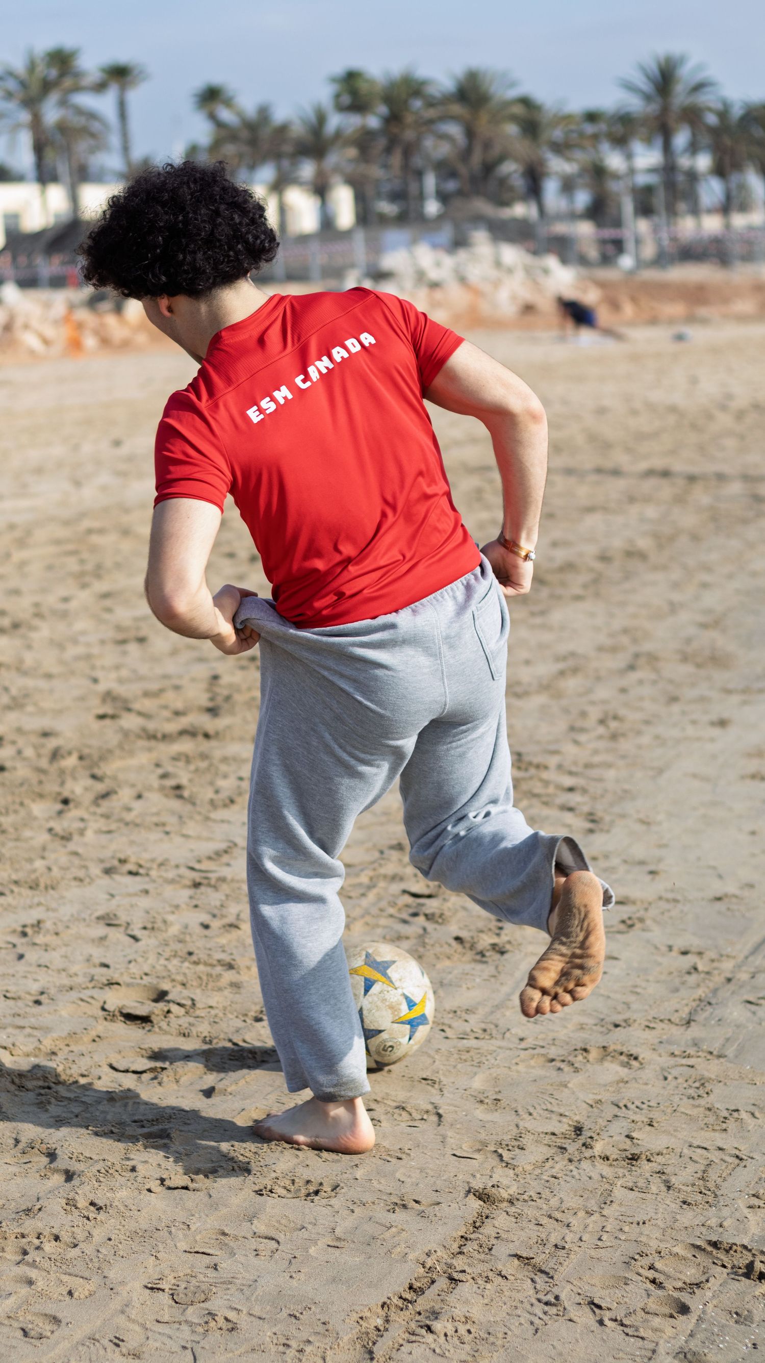 Soccer player in red shirt practicing ball control on sandy beach.