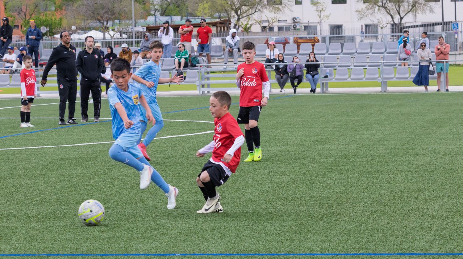 Youth soccer match between teams in red and blue uniforms on artificial turf field.