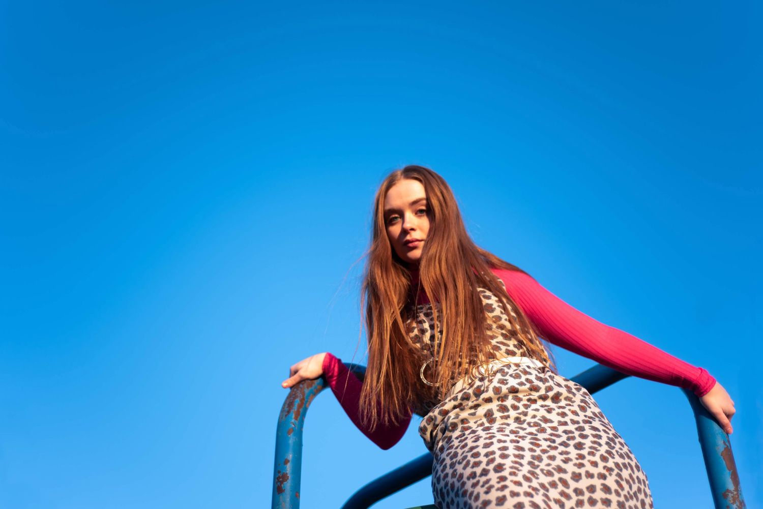 Person in leopard print dress poses against bright blue sky on playground equipment.