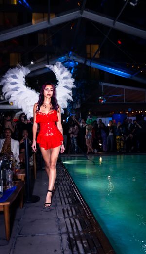 Model walks poolside runway in red bodysuit with dramatic white feather headdress.