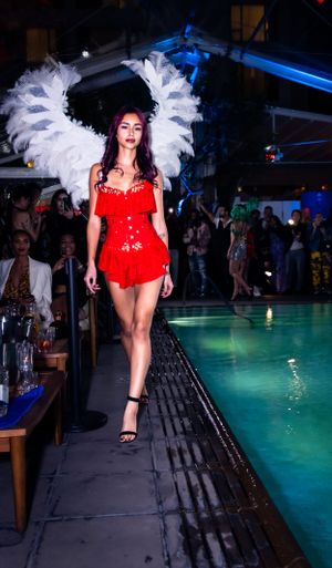 Fashion show features red ensemble with elaborate white feather headpiece against pool backdrop.