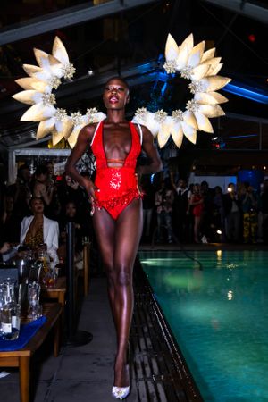 Model in red swimsuit walks runway wearing dramatic gold feathered wing headpiece beside illuminated pool.