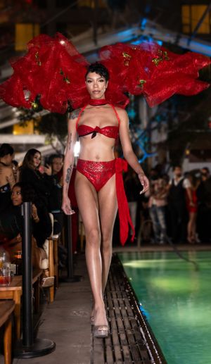 Fashion model in red two-piece ensemble with flowing fabric walks runway beside pool.